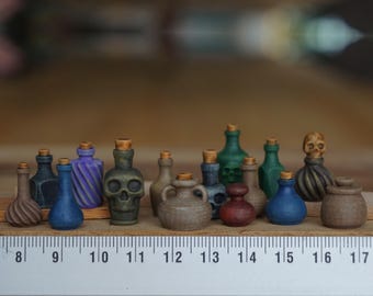 May include: A collection of 15 miniature glass bottles with cork stoppers. The bottles are various colors, including brown, green, blue, purple, and black. Some bottles have a skull design, while others have a swirl design. The bottles are arranged on a wooden surface with a ruler.