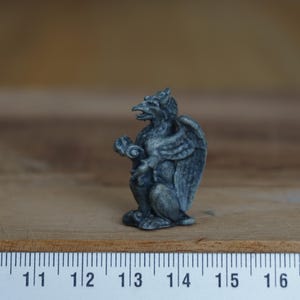Może przedstawiać: Mała, szara, kamienna figurka gargoyli z skrzydłami i zakręconym ogonem. Gargoyle siedzi na drewnianej powierzchni.