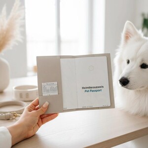 May include: A light beige leather pet passport cover holding an open document with the text Heimtierausweis Pet Passport printed on the white page. The cover features a small interior pocket containing a white card with printed text lines.