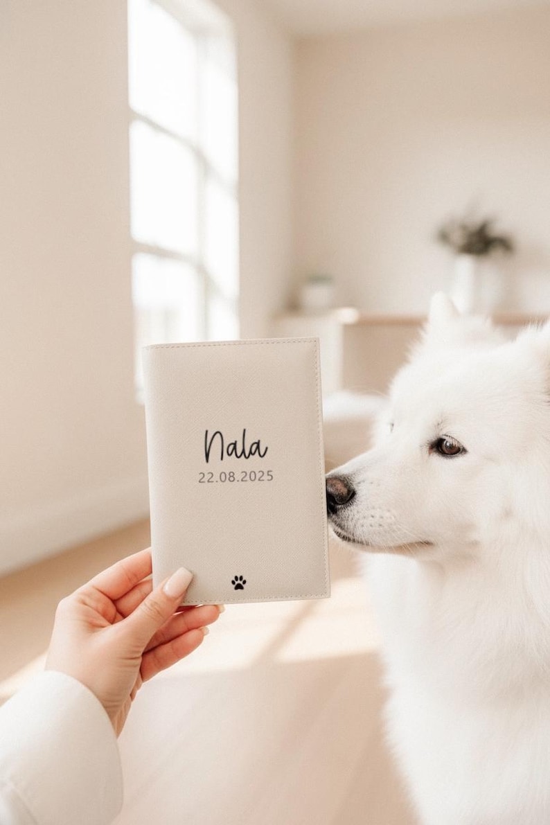 May include: A light beige rectangular passport holder featuring the name Nala in black script, the date 22.08.2025 in black text, and a small black paw print icon centered at the bottom. The cover has a smooth, matte finish and stitched edges.