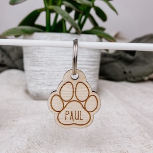 May include: Wooden dog tag with a paw print design and the name "PAUL" engraved on it. The tag is attached to a silver key ring.
