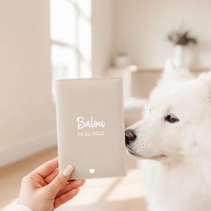 May include: A light beige rectangular passport holder featuring the white printed name Balou, the date 09.02.2022, and a small white heart icon at the bottom center. The cover has a smooth, matte texture with visible stitching along the edges.