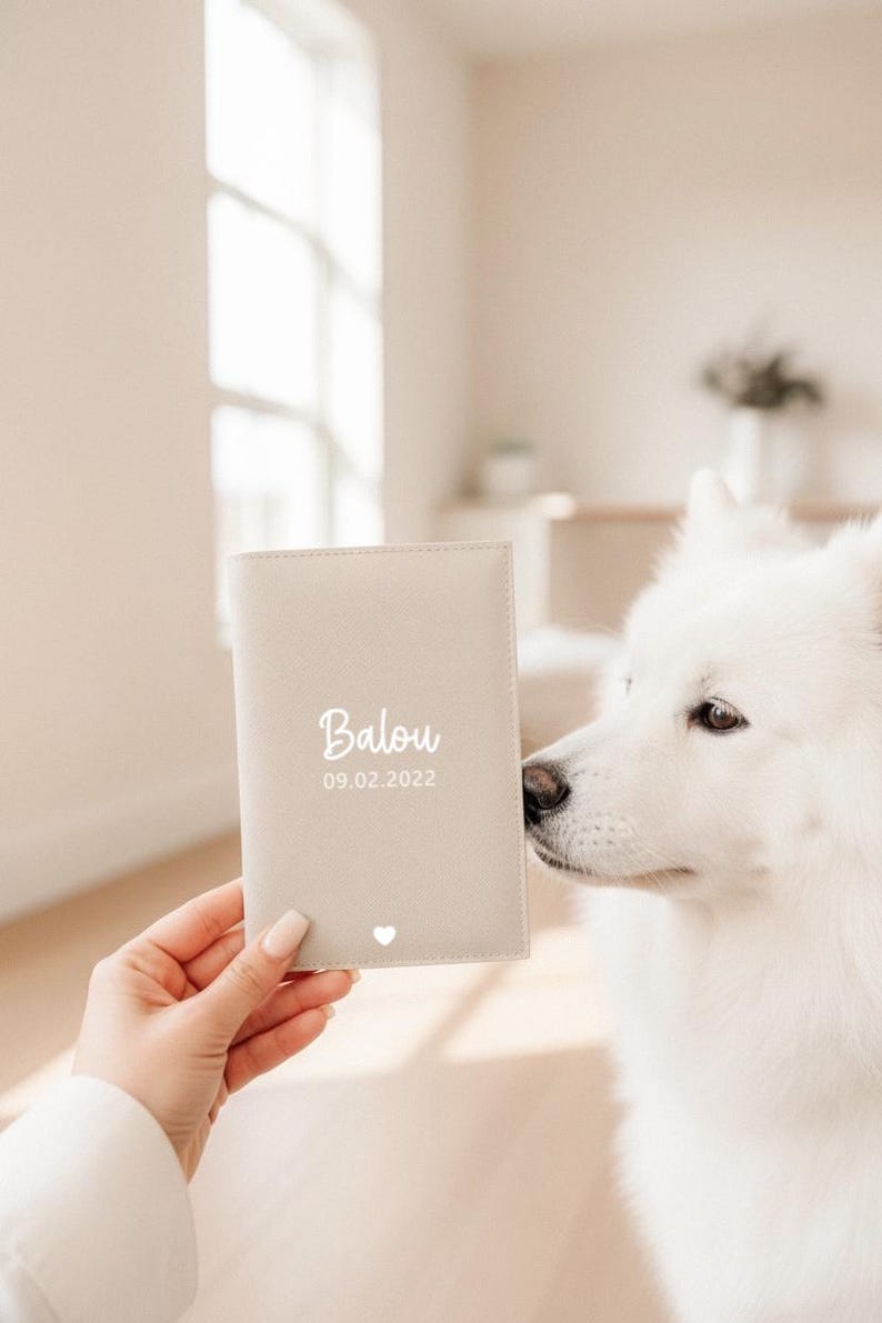 May include: A light beige rectangular passport holder featuring the white printed name Balou, the date 09.02.2022, and a small white heart icon at the bottom center. The cover has a smooth, matte texture with visible stitching along the edges.