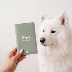 May include: A sage green passport holder featuring the white printed name Freya, the date 22.08.2025, and a small white paw print icon. The rectangular cover has a textured finish and is held upright against a plain, light-colored background.