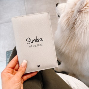 May include: A rectangular, off-white passport holder with a textured finish. The front features the black printed name Simba, the date 07.08.2023, and a small black paw print icon at the bottom center. The item is held against a white, fluffy surface.
