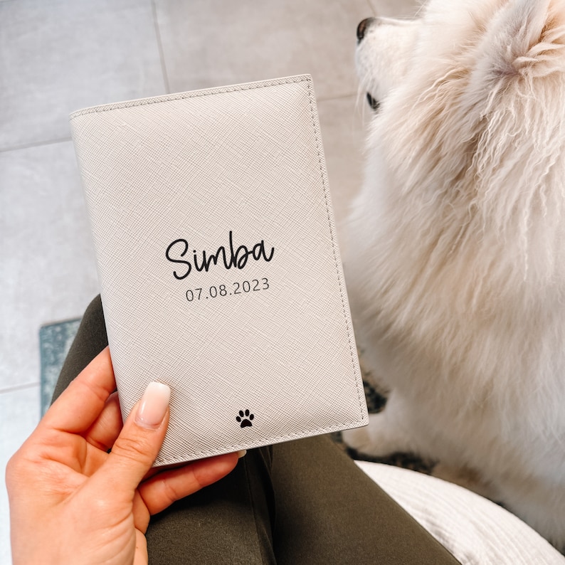 May include: A rectangular, off-white passport holder with a textured finish. The front features the black printed name Simba, the date 07.08.2023, and a small black paw print icon at the bottom center. The item is held against a white, fluffy surface.