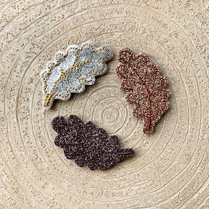 May include: Three glitter oak leaf shapes in silver, copper, and brown. The leaves are arranged in a circle on a wood background.