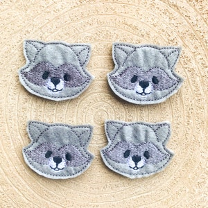 May include: Four gray felt raccoon face appliques with embroidered details. The appliques have black eyes, a black nose, and a white mouth.