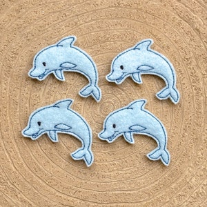 May include: Four light blue felt dolphin appliques with black embroidered eyes and mouths. The dolphins are arranged in a circle on a brown background.
