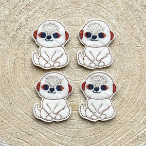 May include: Four white felt appliques of meerkats with brown eyes and orange noses. The meerkats are sitting with their paws crossed and have a simple embroidered design.