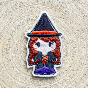 Könnte beinhalten: Gestickter Filz-Hexen-Patch mit einem schwarzen Hut, orangem Rand, braunen Haaren und einem lila Kleid.