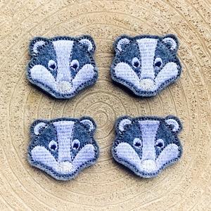 以下が含まれることがあります： 木製の背景に、薄い青色のバジャーの顔が刺繍されたフェルトパッチが4つあります。