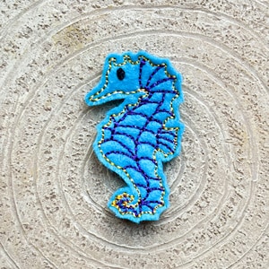 May include: A blue felt seahorse with gold and purple embroidery. The seahorse is standing upright on a light brown background.