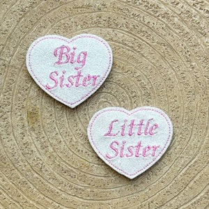 Pode incluir: Dois patches em forma de coração de feltro branco com bordado rosa. O patch superior diz "Big Sister" e o patch inferior diz "Little Sister".