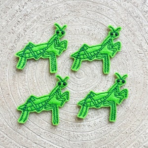 May include: Four green felt embroidered praying mantises with black eyes. The praying mantises are arranged in a circle on a white background.