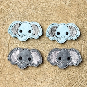 May include: Four felt elephant patches in light blue and grey. Each patch has embroidered details, including ears, eyes, and outline. The patches are arranged on a wooden surface.