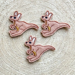 Può includere: Tre applicazioni di canguro in feltro di colore rosa chiaro con dettagli di ricamo bianco e dorato. I canguri sono in posizione di salto con la coda arrotolata.
