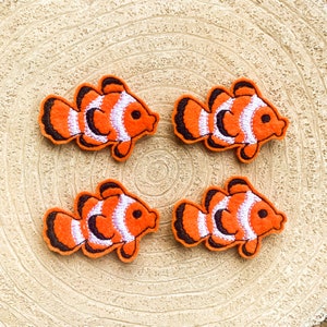 May include: Four orange, white, and black clown fish patches. Each patch features a detailed design of a clown fish with white stripes, black outlines, and a small black eye. The patches are arranged on a light brown, wood-grain surface.