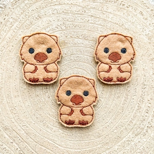 Könnte beinhalten: Drei braune Filzapplikationen eines Cartoon-Wombats mit schwarzen Augen und einer braunen Nase. Die Wombats sitzen mit gekreuzten Armen.