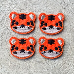 May include: Four orange tiger face patches with black markings and white embroidered details. Each patch has a stitched orange border. The patches are arranged on a light-coloured, textured surface.