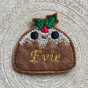 May include: A brown felt Christmas pudding patch with embroidered details. The patch features a smiling face, holly leaves, and the name "Evie" in gold script. The patch is approximately 3 inches wide.