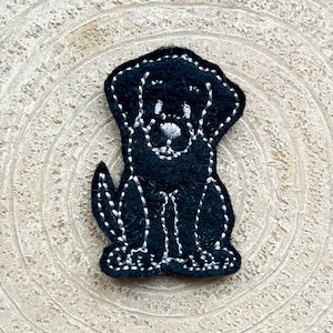 May include: Black felt dog applique with white stitching. The dog is sitting with its legs visible and has a simple face with a nose and eyes.