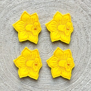 May include: Four yellow felt daffodil flower shapes with embroidered details. The flowers are arranged in a square pattern on a gray background.