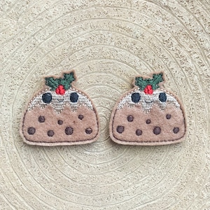 May include: Two brown felt Christmas pudding ornaments with green holly and red berries on top. Each pudding has a smiling face with black eyes and a white mouth.