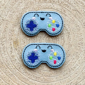 Puede incluir: Dos mandos de juego de fieltro azul bordados con caras felices. Cada mando tiene un botón azul, rojo, verde y amarillo.