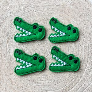 May include: Four green felt alligator head patches with open mouths, white teeth, and black eyes. The patches are arranged on a light brown, textured surface, likely wood.