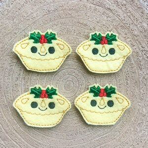 Op de afbeelding: Vier kerstmincepies van vilt met geborduurde hulst- en bessenversieringen en glimlachende gezichten.