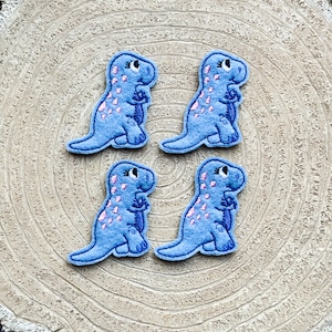 May include: Four blue felt dinosaur appliques with pink accents. The dinosaurs are standing upright with their arms crossed.