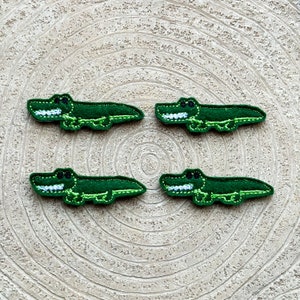 May include: Four green felt alligator appliques with white teeth and eyes. The appliques are on a light brown wood background.