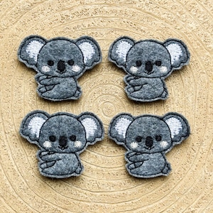 May include: Four gray felt koala appliques with white embroidered ears and faces. The koalas are all facing forward with their arms crossed.