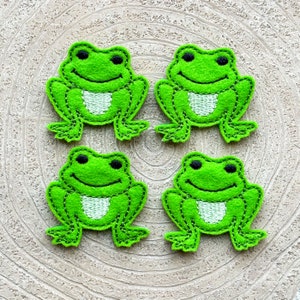 Könnte beinhalten: Vier grüne Filz-Frosch-Applikationen mit weißen gestickten Augen und Mündern. Die Frösche sitzen auf einer hellbraunen Holzoberfläche.