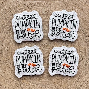 Könnte beinhalten: Vier weiße, bestickte Aufnäher mit glitzernder Textur, die jeweils den schwarzen gestickten Text 'cutest PUMPKIN IN THE patch' und zwei kleine orangefarbene gestickte Herzen zeigen. Die Aufnäher haben einen gewellten Rand.