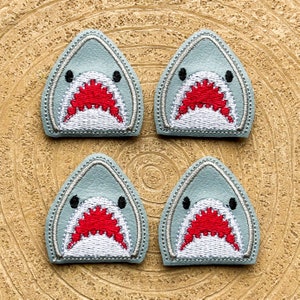 May include: Four grey felt shark patches with white teeth and red mouths. The patches are stitched with white thread and have a white outline.