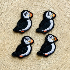 Op de afbeelding: Vier zwarte en witte geborduurde papegaaiduikers met oranje snavels. De papegaaiduikers zijn in een vierkant patroon op een lichtbruin houten oppervlak gerangschikt.