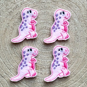 May include: Four pink felt dinosaur appliques with purple spots and pink stitching. Each dinosaur has a pink embroidered eye with a white pupil.
