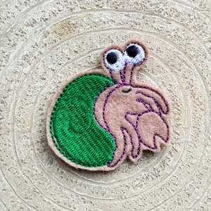 May include: A felt applique of a hermit crab with big eyes. The crab is brown with purple stitching and is carrying a green shell.