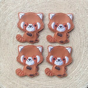 May include: Four embroidered patches featuring a red panda design. Each patch has a brown body, white accents, and a fluffy tail. The patches are arranged in a square pattern on a light brown, textured surface.