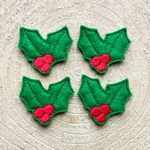 Può includere: Quattro decorazioni in feltro a forma di foglie di agrifoglio con bacche rosse. Le foglie di agrifoglio sono verdi e hanno una texture trapuntata.