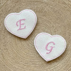 Peut inclure: Deux écussons blancs en forme de cœur avec des détails brodés roses. L'un porte la lettre "E", l'autre la lettre "G". Les bords sont soulignés de coutures roses. Les écussons sont sur une surface texturée brun clair.