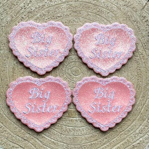 Puede incluir: Cuatro adornos de corazón rosa con purpurina con el texto "Big Sister" en letras blancas.