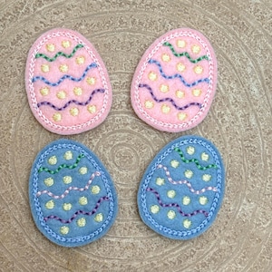 May include: Four felt Easter eggs with embroidered designs. Two eggs are pink with yellow, green, and purple embroidery. Two eggs are blue with yellow, green, and purple embroidery.