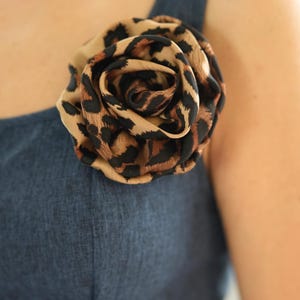 Pode incluir: Uma flor de tecido com estampa de leopardo, em forma de rosa, está presa a uma peça de roupa azul. A flor é feita de tecido bege com manchas pretas. A peça de roupa é de cor azul lisa, criando um contraste.
