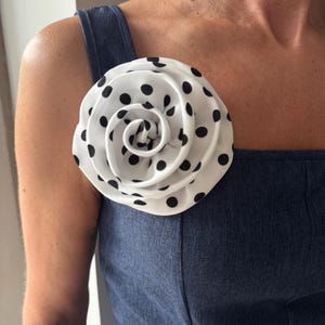 Może przedstawiać: Duży, biały kwiat z czarnej tkaniny w grochy jest przypięty do dżinsowej niebieskiej sukienki. Kwiat ma kształt róży, z płatkami zwiniętymi do wewnątrz. Sukienka ma kwadratowy dekolt i szerokie ramiączka.