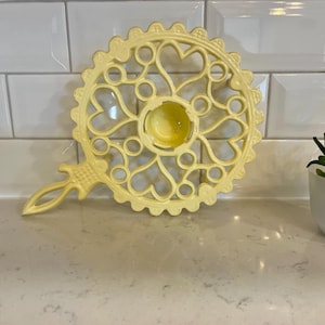 May include: A yellow trivet with a heart and circle design. The trivet has a handle and a central circular area. The trivet is on a white countertop with a small potted plant in the background.