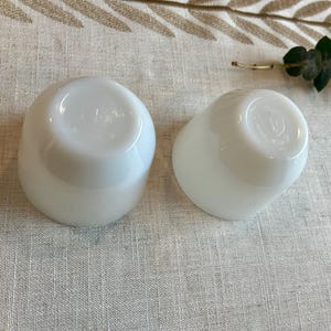 May include: Two white, opaque, rounded glass cups are displayed on a neutral-toned linen surface. The cups have a smooth, glossy finish and a slightly flared shape. The bottoms of the cups are visible, showing a subtle design. A sprig of greenery is in the background.