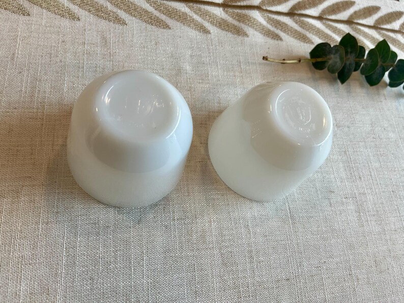 May include: Two white, opaque, rounded glass cups are displayed on a neutral-toned linen surface. The cups have a smooth, glossy finish and a slightly flared shape. The bottoms of the cups are visible, showing a subtle design. A sprig of greenery is in the background.
